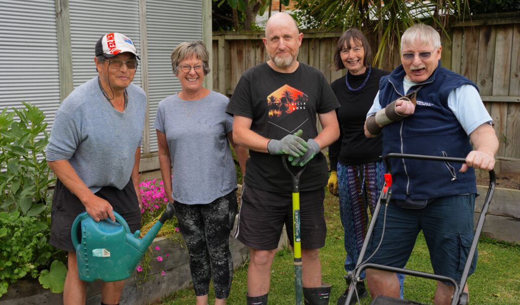 Group of five adults standing in a row in a garden setting smiling at the camera. Some have gardening gear, including a watering can, spade and lawn mower.